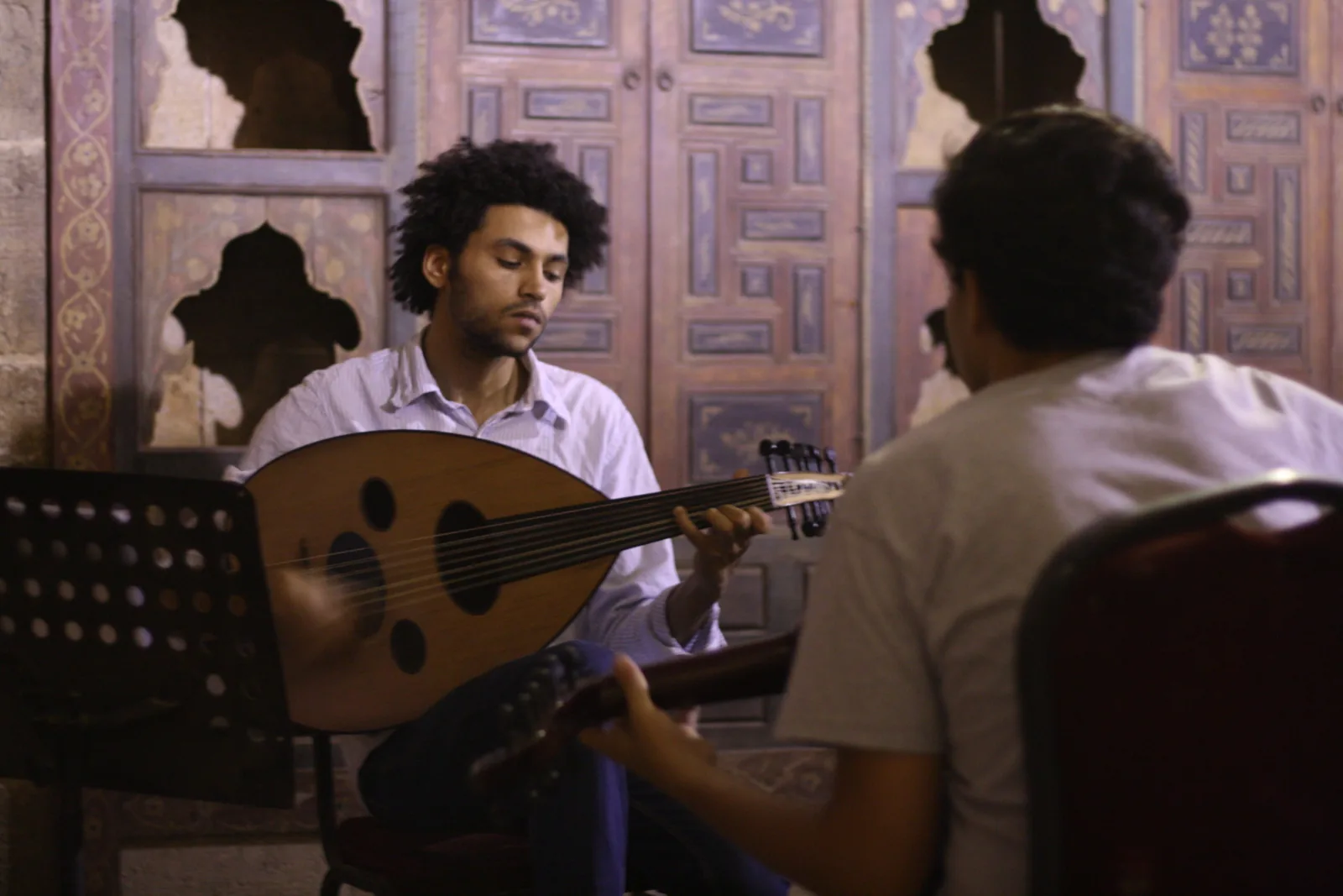 A young man playing an oud, the pear-shaped lute, in front of carved wooden doors, with a second musician in the foreground
